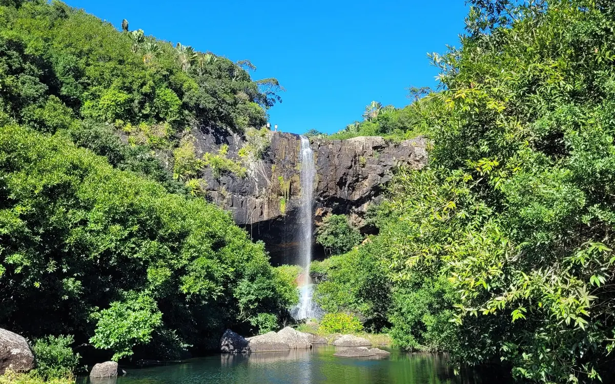 Une grande cascade fine se détachant d&rsquo;une falaise sombre, encadrée d&rsquo;arbres verts denses sous un ciel bleu