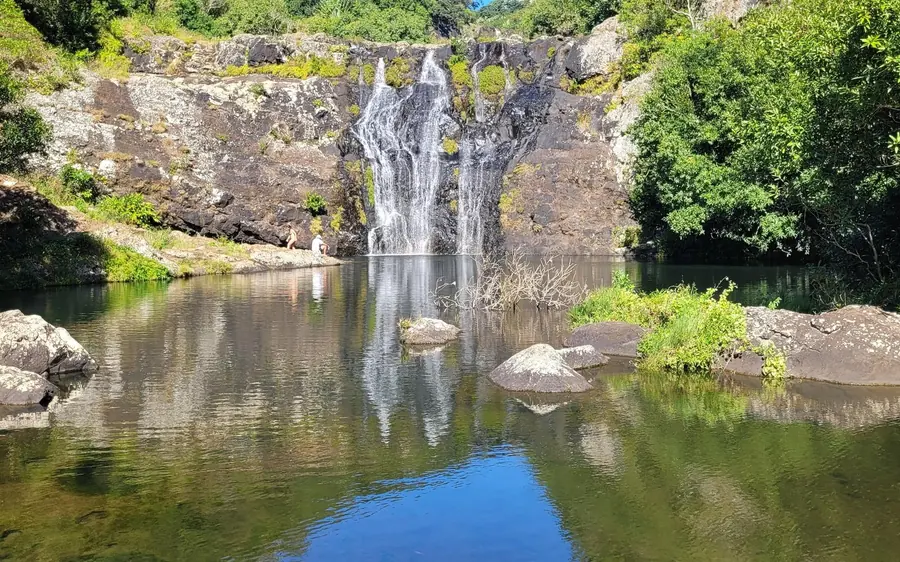 Une large cascade basse sur une paroi rocheuse moussue, un bassin vert immobile et une personne sur la berge