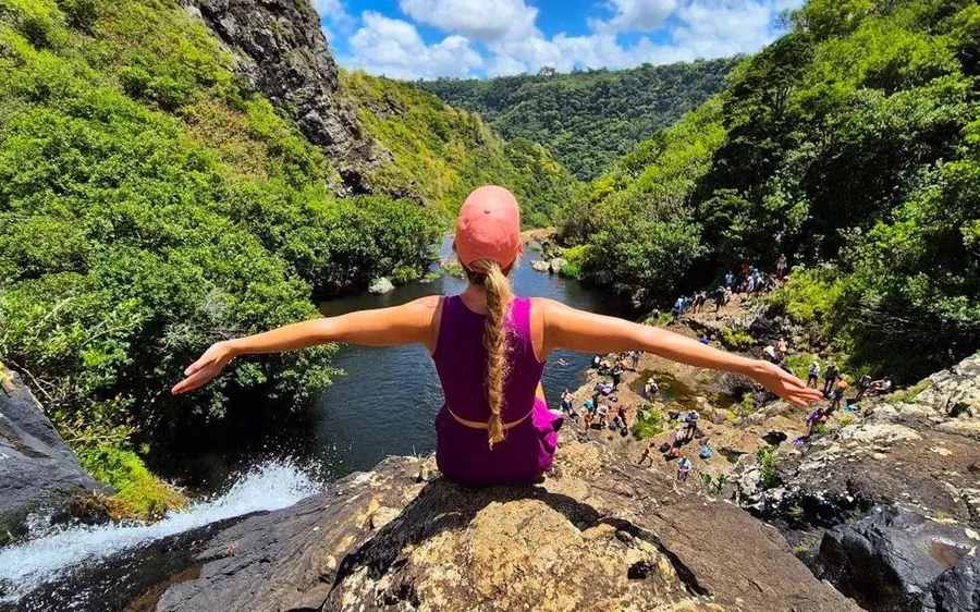 Une femme en haut rose assise sur un rebord rocheux, bras écartés, un bassin de rivière et des personnes en contrebas