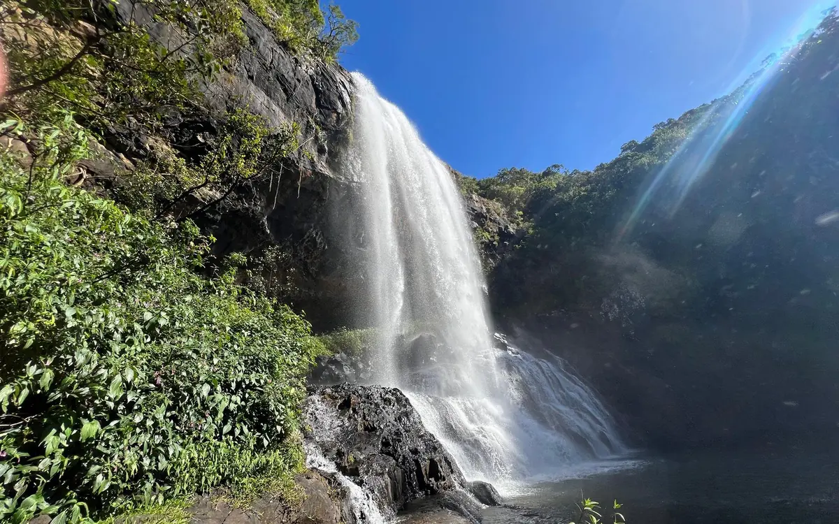 Une large cascade tombant d'une roche sombre dans un bassin sombre, avec une végétation dense et verte de part et d'autre